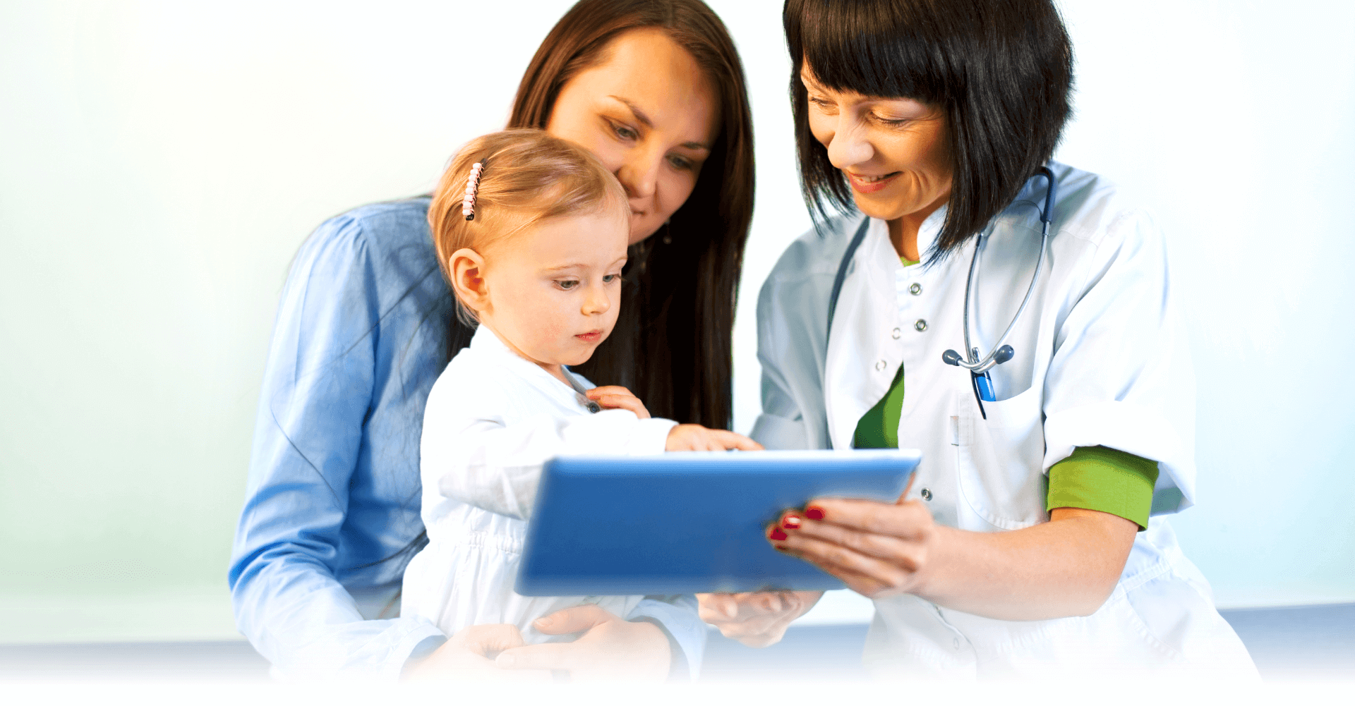 Pediatrician shows a girl toddler cradled in her mother’s lap her health records with a tablet.